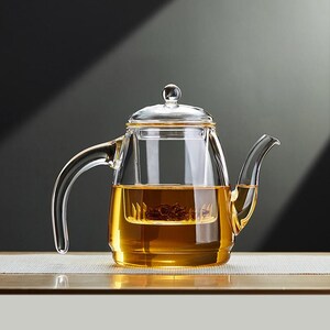 Könnte beinhalten: Eine transparente Glas-Teekanne mit gebogenem Griff und Ausguss. Die Teekanne ist mit bernsteinfarbenem Tee gefüllt und hat einen eingebauten Infuser. Der Deckel hat einen kleinen, runden Knopf. Die Teekanne steht auf einer hellen Oberfläche vor dunklem Hintergrund.