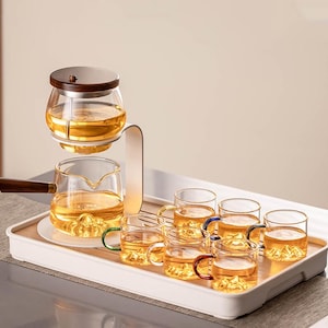 Puede incluir: Un juego de té de vidrio con asa y tapa de madera, que incluye una tetera y varias tazas pequeñas. Las tazas tienen asas de colores y están llenas de té. El juego está dispuesto sobre una bandeja blanca.