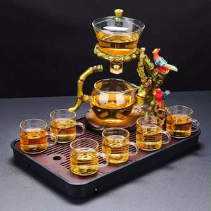May include: A glass tea set with a teapot, a serving cup, and six small teacups, all filled with tea. The set includes a decorative bamboo-style stand with colorful bird figurines. The tea set is on a dark tray.