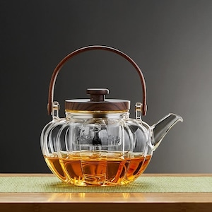 Op de afbeelding: Een heldere glazen theepot met een houten handvat en deksel. De theepot heeft een afgeronde vorm met verticale ribbels en is gevuld met amberkleurige vloeistof. De handgreep is gebogen en van donker hout.