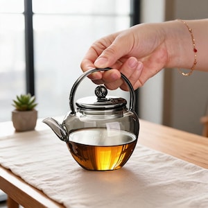 May include: A clear, smoke-tinted glass teapot with a rounded body and a matching lid featuring a small, dark spherical knob. The teapot has a curved handle and a short spout, and it is filled with a golden-brown liquid tea.