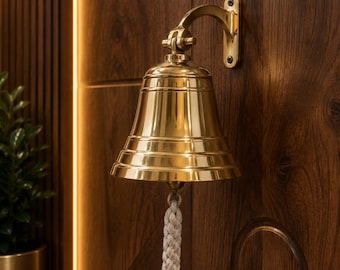 May include: A brass bell with a braided rope pull is mounted on a dark wood door. The bell has a traditional shape with a flared base and a curved bracket. The door is made of dark wood with a rich, warm colour.