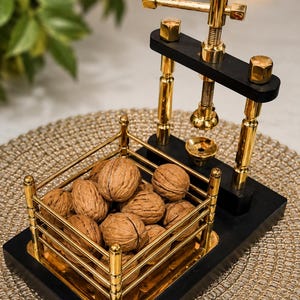 May include: A mechanical nutcracker featuring a black wooden base, gold-toned metal vertical supports, and a screw-driven pressing mechanism. A square gold-toned metal basket with vertical railings holds a collection of whole brown walnuts.