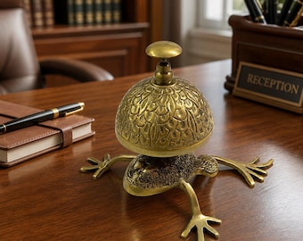 May include: A brass-coloured metal desk bell featuring a frog-shaped base with four extended legs and a textured, dome-shaped top with a knob handle. The bell has a detailed, scale-like pattern on the dome and a dark, mottled finish on the frog body.