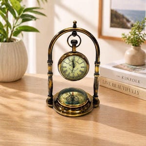 May include: A vintage-style brass desk clock with a compass. The clock face is cream with black Roman numerals and hands. The clock is suspended in an arched frame. The base features a compass with a glass cover. The clock measures approximately 15 cm in height.