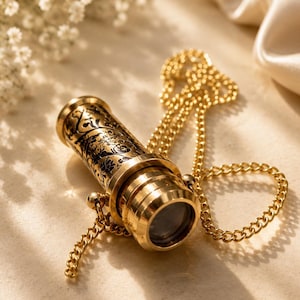 May include: A gold-toned brass monocular pendant featuring an intricate black floral engraving on the cylindrical body. The optical instrument is attached to a matching gold-coloured metal chain, showcasing a polished finish and a circular lens.