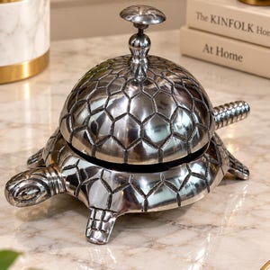 May include: A silver-toned metal desk bell shaped like a turtle. The shell of the turtle forms the dome of the bell, featuring a textured, hexagonal pattern. A small, round button sits at the top of the dome, and a tail extends from the back.