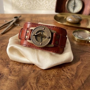 May include: A wide brown leather cuff bracelet featuring a circular brass sundial centrepiece. The sundial has engraved markings and a central gnomon. The leather band is secured with brass rivets on both sides of the circular metal component.