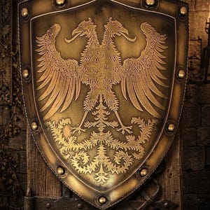 Puede incluir: Un escudo con acabado en bronce metálico que presenta un relieve de un águila bicéfala. El escudo tiene una base puntiaguda y está decorado con una serie de remaches circulares a lo largo del borde exterior, creando un aspecto estructurado.