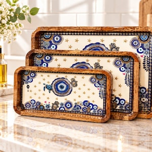 May include: A set of three rectangular wooden serving trays with rounded corners. The interior surfaces feature a cream background decorated with blue and gold evil eye motifs, small gold stars, and turtle illustrations, framed by a dark wood border.