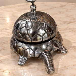 May include: A silver-toned metal desk bell shaped like a turtle. The shell forms the dome of the bell, featuring a textured, geometric hexagonal pattern. A small, rounded knob sits at the top, and the turtle's head and feet extend from the base.