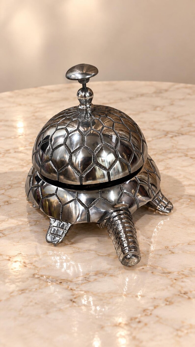 May include: A silver-toned metal desk bell shaped like a turtle. The shell forms the dome of the bell, featuring a textured, geometric hexagonal pattern. A small, rounded knob sits at the top, and the turtle's head and feet extend from the base.