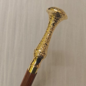 May include: A walking stick featuring a polished gold-tone metal handle with an intricate embossed leaf pattern. The handle is shaped with a flared top and connects to a smooth, dark brown wooden shaft that extends downward from the metal grip.