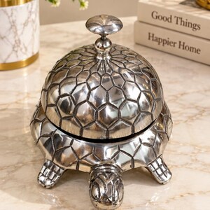 May include: A silver-toned metal desk bell shaped like a turtle. The bell features a textured, geometric shell pattern that covers the dome and the base. It has a small, rounded handle at the top and a protruding turtle head and four feet.