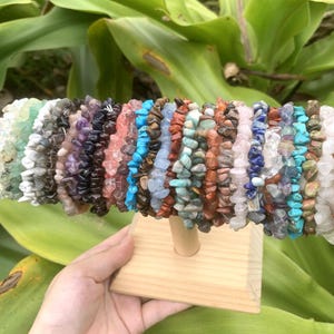 May include: A display of gemstone chip bracelets in various colors, including green, purple, blue, and brown. The bracelets are made of small, irregularly shaped stones and are displayed on a wooden stand. The background features green leaves.