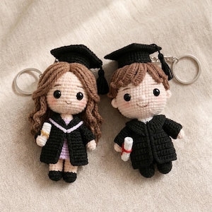 Puede incluir: Dos llaveros de graduación de ganchillo. Cada muñeca lleva una toga y un birrete negros. Una muñeca tiene el pelo castaño y la otra castaño claro. Ambas muñecas sostienen un diploma en miniatura. Fondo neutro.