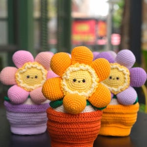 Könnte beinhalten: Drei gehäkelte Blumentopfdekorationen. Jede Blume hat ein lächelndes Gesicht und bunte Blütenblätter in Rosa-, Orange- und Lilatönen. Die Töpfe sind orange, lila und gelb. Die Blumen stehen auf einer schwarzen Oberfläche.