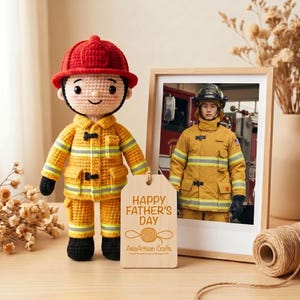May include: A handmade crochet firefighter doll wearing a red helmet and yellow uniform, standing next to a framed photo of a firefighter. A wooden tag reads "Happy Father's Day".