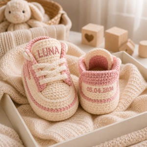 Puede incluir: Un par de zapatillas de bebé tejidas a ganchillo en color crema y rosa. La zapatilla izquierda tiene el nombre LUNA bordado en la lengüeta. La derecha tiene la fecha 05.04.2024 bordada en el talón. Ambos zapatos tienen cordones blancos.