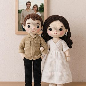 Könnte beinhalten: Zwei handgefertigte Häkelpuppen, ein Paar, stehen nebeneinander. Die männliche Puppe trägt ein beiges Hemd und eine schwarze Hose, während die weibliche Puppe ein weißes Kleid trägt. Im Hintergrund hängt ein gerahmtes Foto eines Paares.