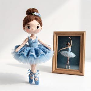 May include: A crocheted ballerina doll with brown yarn hair in a bun, wearing a blue leotard and a layered tulle tutu. The doll has blue ballet slippers with ribbon ties. Beside the doll is a wooden picture frame containing a photo of a ballerina.