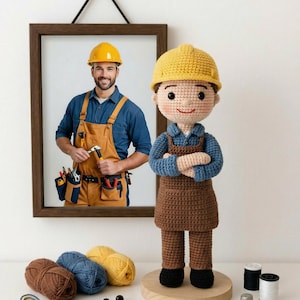 May include: A framed photo of a construction worker hangs above a crocheted doll of a construction worker. The doll wears a yellow hard hat, blue shirt, and brown overalls. Yarn, beads, and sewing notions are on the table.