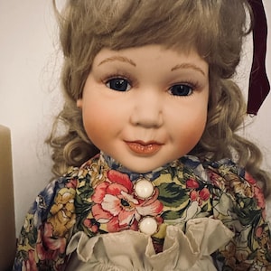May include: A porcelain doll with blonde, curly hair and blue eyes. The doll is dressed in a floral print dress with large white buttons and a ruffled collar. A burgundy bow is in the doll's hair. The doll has rosy cheeks and a gentle smile.