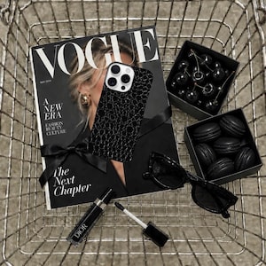 Può includere: Una cover per smartphone nera con motivo a coccodrillo testurizzato, una rivista Vogue, una scatola di ciliegie scure, una scatola di macaron neri, occhiali da sole neri e un lucidalabbra Dior con applicatore in un cestino di metallo.