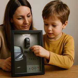 May include: A framed Tesla Model Y art piece. The black frame holds a gray background with a Tesla car illustration, a miniature car, and the word "TESLA". The frame is held by a person and a child.