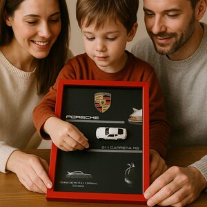 Könnte beinhalten: Ein rot gerahmtes Display mit schwarzem Hintergrund, das ein Porsche-Logo und ein weißes 911 Carrera RS Modellauto zeigt. Das Display enthält den Text "PORSCHE" und Fahrzeugspezifikationen. Eine Familie betrachtet das Display.