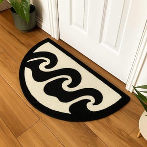 Könnte beinhalten: Eine halbkreisförmige Fußmatte mit schwarzem Rand und cremefarbenem Innenraum. Der cremefarbene Innenraum weist ein schwarzes, wellenartiges Design auf. Die Matte liegt vor einer weißen Tür auf einem Holzboden. Eine Topfpflanze ist im Hintergrund zu sehen.