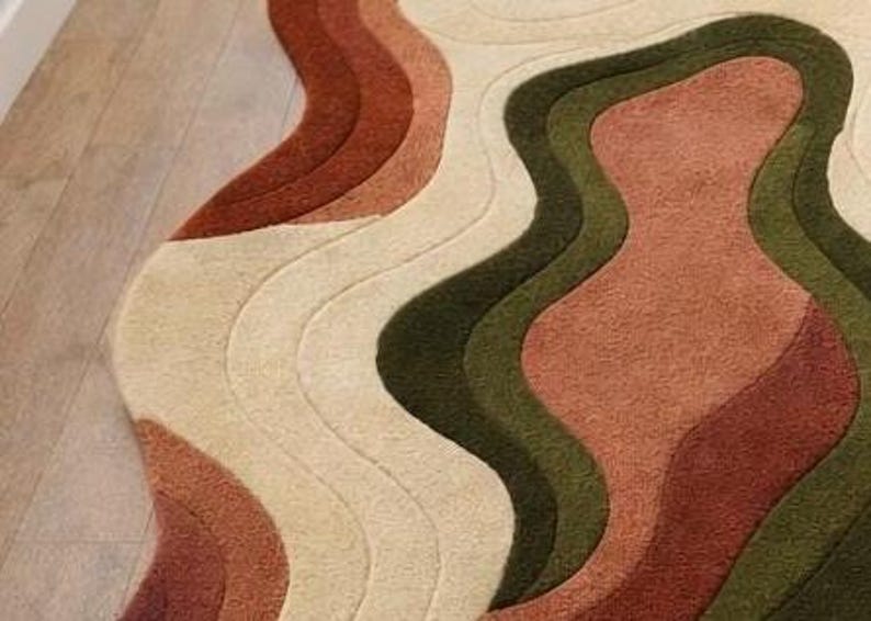 Pode incluir: Um close-up de um tapete com um design ondulado e em camadas. O tapete apresenta tons de bege, castanho e verde, criando um padr&atilde;o abstrato texturizado. O design do tapete assemelha-se a um mapa topogr&aacute;fico ou a uma paisagem estilizada.