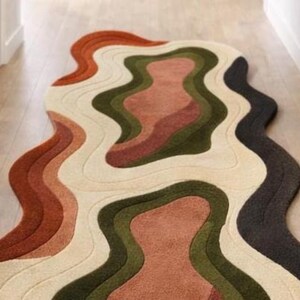 Pode incluir: Um tapete abstrato com um design ondulado e em camadas. A paleta de cores do tapete inclui ferrugem, creme, verde-oliva e cinza carv&atilde;o. &Eacute; colocado sobre um piso de madeira clara, adicionando um toque moderno ao espa&ccedil;o. Medidas em cent&iacute;metros.