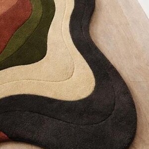 Pode incluir: Um tapete moderno e abstrato com um design ondulado. O tapete apresenta uma paleta de cores castanho, verde-oliva, creme e cinzento escuro. O tapete &eacute; feito de um material macio e texturizado e est&aacute; colocado num ch&atilde;o de madeira.