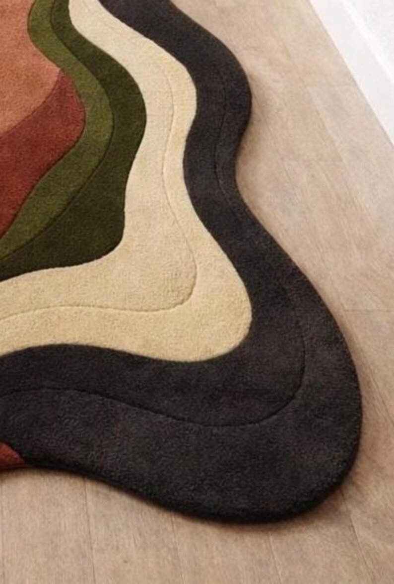 Pode incluir: Um tapete moderno e abstrato com um design ondulado. O tapete apresenta uma paleta de cores castanho, verde-oliva, creme e cinzento escuro. O tapete &eacute; feito de um material macio e texturizado e est&aacute; colocado num ch&atilde;o de madeira.