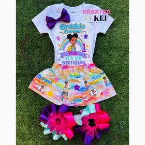May include: A white t-shirt and a colorful skirt set with matching hair bows. The shirt features the text "Gracie's Corner" and "It's My Birthday". The skirt has a rainbow design with cartoon characters. Purple bow.