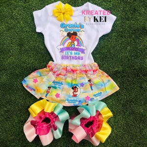 May include: A white baby bodysuit with a yellow bow, a colorful skirt, and two matching hair bows. The bodysuit has a graphic that says "Gracie's Corner" and "It's My Birthday". The skirt and bows have a rainbow design.