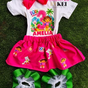 May include: A white t-shirt with a Dora the Explorer graphic, the number 3, and the name Amelia, paired with a hot pink skirt featuring Dora and Boots characters, a large hot pink hair bow, and two green and white ruffled leg warmers.