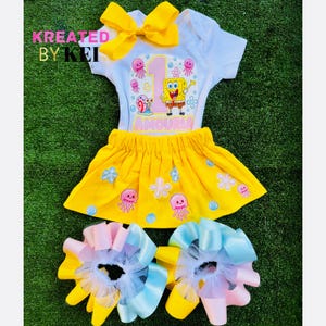 May include: A yellow and white outfit for a child, featuring a white top with a SpongeBob SquarePants graphic and a yellow skirt. The outfit includes matching hair bows with yellow, pink, and blue ribbons. The text "KREATED BY KEI" is visible.