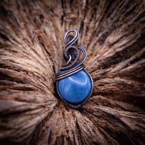 Könnte beinhalten: Ein handgefertigter Anhänger mit einem hellblauen, tropfenförmigen Edelstein, der kunstvoll in dunklen, oxidierten Metalldraht gewickelt ist. Das Design des Anhängers kontrastiert mit dem glatten, polierten Stein. Er wird auf einer strukturierten, braunen Holzoberfläche präsentiert.