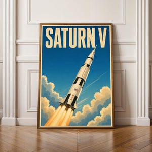 Puede incluir: Un póster vertical que muestra un cohete Saturn V despegando hacia un cielo azul con nubes. El cohete es blanco con marcas negras, el texto 'SATURN V' en letras grandes de color crema en la parte superior, y 'USA' impreso en el lateral con una pequeña bandera estadounidense.