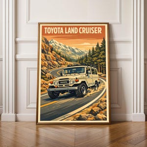 以下が含まれることがあります： 山道を走るヴィンテージトヨタランドクルーザーを描いたフレームプリント。レトロな旅行ポスター風で、上部に「TOYOTA LAND CRUISER」の文字。車両はクリーム色です。