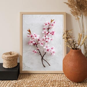 May include: A framed print featuring a close-up of a cherry blossom branch with delicate pink flowers and brown stems. The artwork is set against a textured white background and framed in light wood. A decorative vase and basket are also in the image.