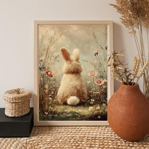 Op de afbeelding: Een ingelijste print met een pluizig, lichtbruin konijn van achteren, zittend in een veld met wilde bloemen. Het kunstwerk heeft een zacht, pastelkleurig kleurenpalet met roze en groene accenten, waardoor een grillige, door de natuur geïnspireerde scène ontstaat.