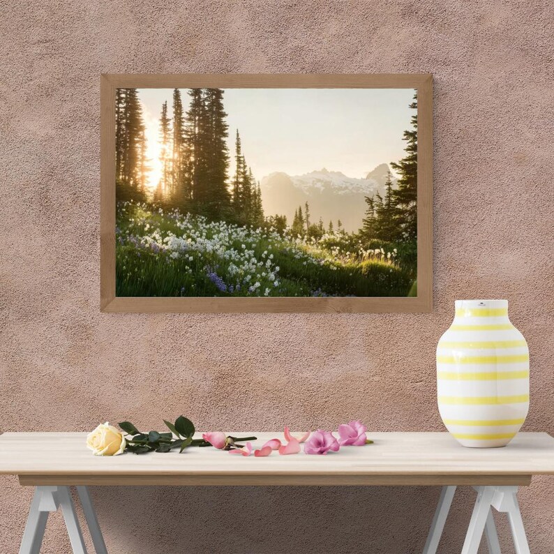 May include: A framed landscape photograph of a mountain scene with a field of wildflowers and tall trees. The sun shines through the trees. A yellow and white striped vase, a yellow rose, and pink flowers are on a table.