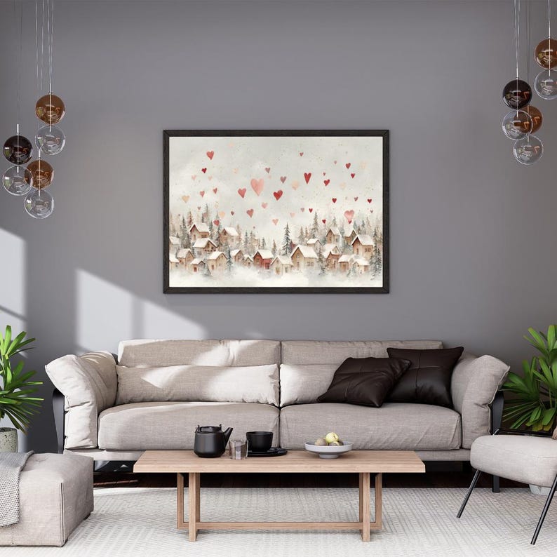 May include: A framed watercolor painting depicts a snow-covered village with red-roofed houses and heart-shaped accents. The artwork is set against a soft, neutral background, creating a cozy, romantic atmosphere. The painting is displayed above a beige sofa.
