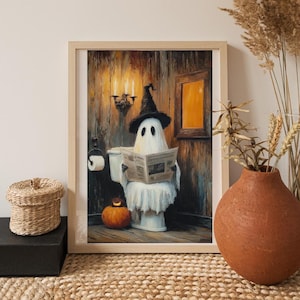 Op de afbeelding: Een ingelijste print toont een spook met een heksenhoed die een krant leest op een toilet. De afbeelding bevat een pompoen, kaarsen en een rustieke badkameromgeving, perfect voor Halloween-decoraties.