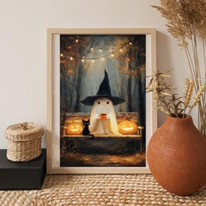 Op de afbeelding: Een ingelijste Halloween-print met een vriendelijke geest die een heksenhoed draagt, een kopje vasthoudt en op een bank zit tussen twee verlichte pompoenen. Een zwarte kat zit in de buurt. De achtergrond toont een bos met lichtslingers.