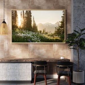 May include: A framed landscape photograph of a mountain scene. The image features a vibrant meadow of white and purple wildflowers, with tall evergreen trees and snow-capped mountains in the background. The sun shines through the trees.