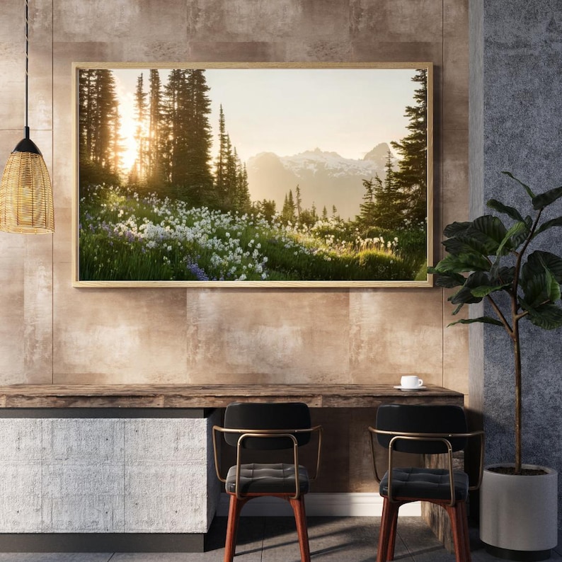 May include: A framed landscape photograph of a mountain scene. The image features a vibrant meadow of white and purple wildflowers, with tall evergreen trees and snow-capped mountains in the background. The sun shines through the trees.